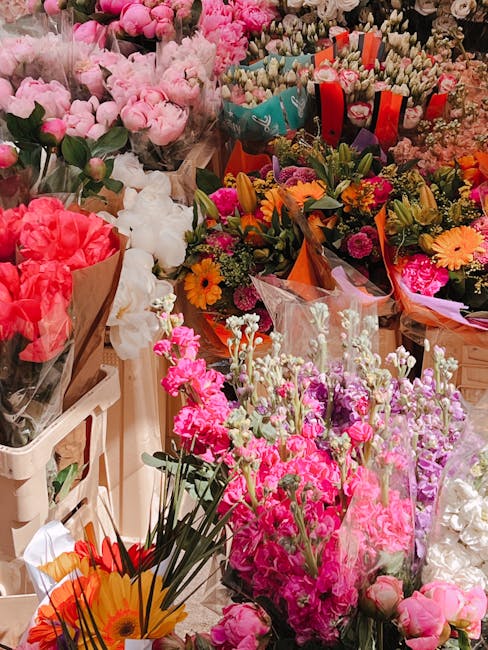 A vibrant display of fresh flowers arranged in various bouquets and wrapped in colorful paper and plastic, featuring a mix of pink, white, red, orange, and purple blooms. Prominent flower types includ