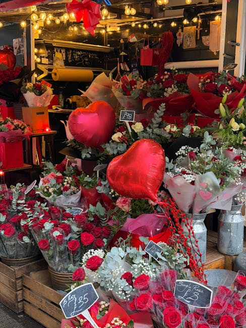 A vibrant display of fresh floral arrangements at a flower stall, featuring predominantly red and white roses, accented with greenery and baby's breath. The bouquet arrangements are wrapped in pink an